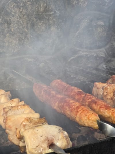 The media file shows a close-up, overhead view of food being grilled outdoors. The subjects are pieces of meat cooking on a grill. There are long, cylindrical sausages and pieces of what appear to be chicken or pork belly. Tongs are visible, holding and manipulating one of the sausages. Smoke is rising from the grill, obscuring parts of the scene. The background suggests a rustic setting, with rough, dark stone or earth visible. The lighting indicates it is daytime. The overall impression is of a barbecue or outdoor cooking event. There is no visible text. The location context is Pidhorodne, Ukraine, suggesting a possible outdoor gathering or a meal prepared in a rural or semi-rural setting in that region.