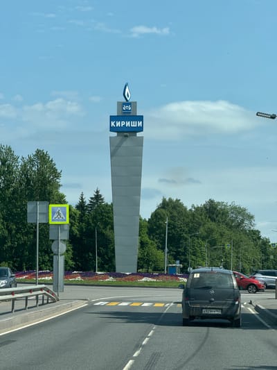 The image depicts a roadside view in the city of Kirishi, Russia, with a prominent monument and a street scene. The monument, a tall, grey, trapezoidal structure, is topped with a blue sign displaying the word "КИРИШИ" in large white letters and a blue flame-like logo above it. The monument stands against a backdrop of a blue sky with scattered white clouds.
In the foreground, a grey asphalt road curves gently. Yellow and white road markings are visible, including a pedestrian crossing. Several cars are present: a dark-colored minivan with a license plate reading "A929PX 147" is in the center of the frame, and other vehicles are partially visible in the distance. A metal guardrail runs along the edge of the road.
To the left of the road, a pedestrian crossing sign with a blue background and a white pedestrian silhouette is mounted on a pole, alongside other traffic signs. Behind the signs, lush green trees form a dense background.
A landscaped area with colorful flower beds featuring red, white, and purple blooms is situated behind the guardrail and between the road and the trees. Streetlights are also visible among the trees. The overall scene suggests a sunny day in an urban or suburban environment.