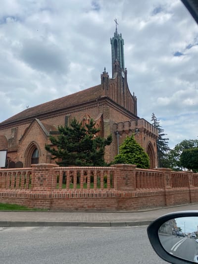 This media file shows a view of a brick church with a green steeple and a cross, taken from a car. The church has arched windows and decorative brickwork. A brick fence is visible in the foreground. The sky is cloudy. In the car's side mirror, other cars and buildings are reflected. The scene appears to be in Mi Miłosław, Poland. The time of day is likely daytime, given the brightness. The weather is overcast.