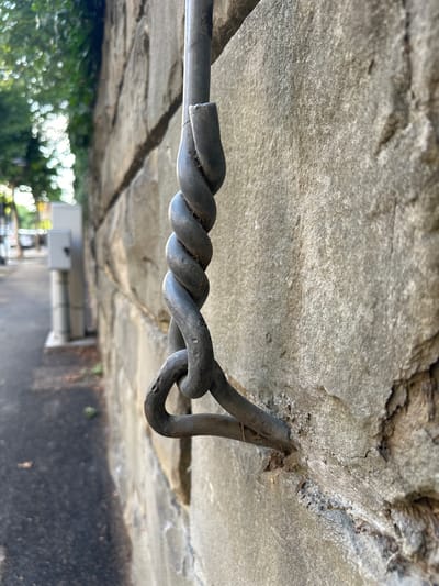 This is a close-up, eye-level, exterior, daylight shot of a weathered, dark grey metal chain link that is attached to a rough, beige stone wall. The chain is twisted and appears to be made of thick, solid metal rods. The stone wall is constructed of large, irregular blocks, showing signs of age and wear. To the left of the wall, a paved pathway is visible, with a faint suggestion of green foliage and trees in the background. The overall scene is devoid of people. The perspective suggests the chain is a functional element, possibly for a barrier or support, embedded into the wall. The location is identified as being in the city of Trieste, Italy.