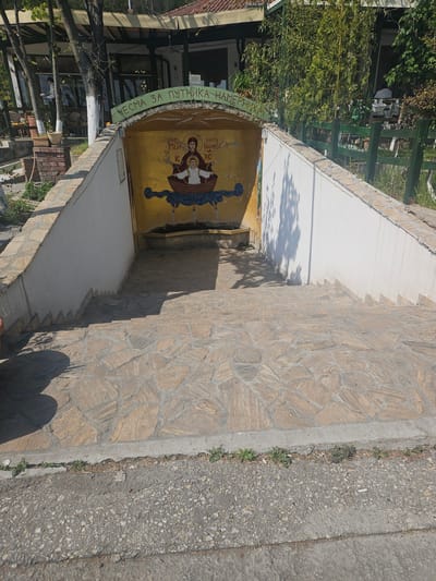 This image captures an outdoor scene in Velika Plana Municipality, Serbia, showing a stone-paved pathway leading down towards an arched entrance. The scene is brightly lit by the sun, casting distinct shadows on the ground and walls, suggesting a clear day in the morning or afternoon.
The foreground features a textured stone pathway on the right, made of irregularly shaped light brown stones. Adjacent to this, on the left, is a concrete border separating it from a darker, asphalt-like surface where small weeds grow in the cracks. A discarded cigarette butt is visible on the darker surface.
The focal point is a white-walled structure with a curved archway, adorned with a vibrant yellow background and religious iconography. This artwork depicts two haloed figures, presumably saints, in a boat sailing on stylized blue waves. Cyrillic text identifies the left figure as "СВЕТИ ПЕТАР" (Saint Peter) and the right figure, holding a book, as "СВЕТИ ЈОВАН БОГОСЛОВ ХС" (Saint John the Theologian HS/Christ). Above the arch, a green band carries the Cyrillic inscription "ЧЕСМА ЗА ПУТНИКА НАМЕРНИКА," which translates to "FOUNTAIN FOR TRAVELERS / WAYFARERS."
Beyond the archway, obscured by dense green foliage and trees, parts of a building with large windows and dark framing are visible, suggesting a restaurant, cafe, or guesthouse. A green wooden trellis or fence can be seen on the far left, adding to the garden-like atmosphere. No people are visible, and the scene is static.