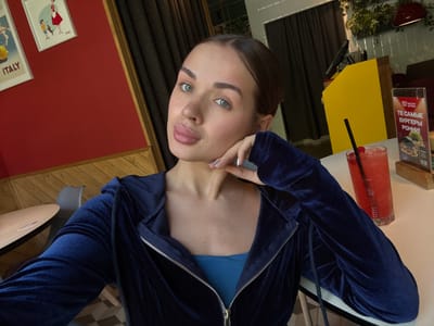 A woman with light skin and light blue eyes is seated at a table, wearing a partially unzipped dark blue velvet tracksuit over a blue sports bra. She has dark hair pulled back from her face. Her left hand is raised towards her face. A red drink with a black straw is on the table to her left, along with a menu stand with text in Russian. Behind her, there is a red wall with framed artwork that includes the word "Italy". The setting appears to be a restaurant or cafe. Based on the location, the city of Borok, Russia can be identified.