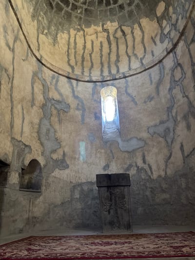 A dimly lit, domed room features aged walls with visible repairs. A small, bright window pierces the wall, casting light. A carved, dark wooden panel displaying a cross is affixed to the wall, adjacent to the edge of a patterned carpet. The room’s architecture suggests an ancient religious structure.