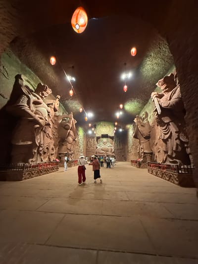 The image captures a view inside the Shizhongshan Grottoes in Shizhong, China. The scene depicts a long cave corridor with stone carvings along the walls. The carvings depict figures, possibly deities or historical individuals, and are illuminated by a series of red lanterns and overhead lights. Tourists are walking along the corridor, observing the carvings. The walls of the corridor appear to be raw rock. The floor is tiled, giving a smooth surface for walking.