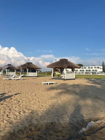 The image depicts a sandy beach with several thatched-roof gazebos. In the background, a modern white building complex is visible, with some trees and communication towers. The sky is blue with scattered white clouds. There are no people in the image. The scene is set in the city of Svitiaz, Ukraine.
