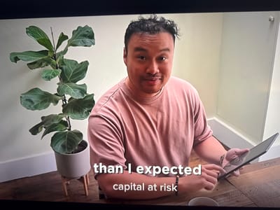 A man is sitting at a wooden table and looking at the camera. He is wearing a pink t-shirt and has curly dark hair. He is holding a black tablet in his left hand and his right index finger is touching the screen. There is a potted plant to his left and a white bowl in the bottom right corner of the image. The background is a plain wall. Subtitles at the bottom of the image read "than I expected" and "capital at risk". There is no discernible information in the image that places it in Cheltenham, United Kingdom.