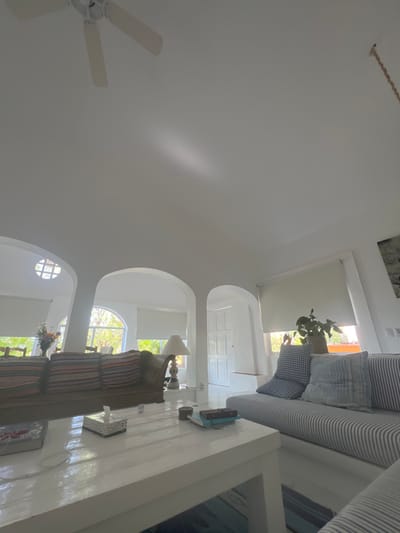 An interior shot captures a room with white walls and arched openings. A striped sofa with blue cushions is positioned near a window with a roller shade. A long white table is in the foreground, upon which several objects rest. A lamp sits on a sofa with striped cushions. Light streams through the windows. A ceiling fan is visible in the upper left corner.
