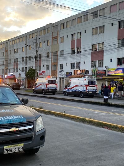 The image depicts a street scene in Quito, Ecuador, characterized by a long, multi-story residential and commercial building. In the foreground, the front portion of a dark-colored transit vehicle is visible, bearing the license plate "PMA-3189" and "ECUADOR" written on it. The vehicle has "TRANSITO" emblazoned on its hood.
On the street, two white ambulances with red and blue checkered patterns are parked. Both ambulances have "AMBULANCIA" written on their sides. One ambulance has its emergency lights illuminated. Several people are gathered on the sidewalk in front of the businesses located in the building. The businesses include establishments with signs such as "CARNES POLLOS," "PELUQUERIA CANINA," "CERVECERIA," "COFFEE," and "SABOR VENEZOLANO." Overhead power lines crisscross the scene. The sky above is cloudy with hints of sunset colors. The overall environment suggests an urban setting with ongoing activity.