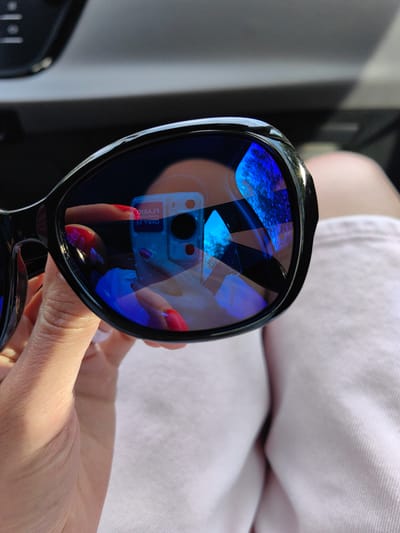 A close-up shot shows a pair of black-rimmed sunglasses being held by a hand with bright red nail polish. The sunglasses' lenses reflect a blue and white digital camera and a hint of the sky. The background is blurred, suggesting an outdoor setting, possibly in the vicinity of Юзуфоўскі сельскі Савет, Belarus, given the specific location context provided.