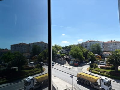 The image depicts a bright, sunny day in Bahçelievler, Turkey, as seen through a large window. The scene outside includes a street with several vehicles, including two yellow-dumping trucks, one white van, a red truck, and several cars. Trees and buildings line the street, with some buildings appearing to be under construction or renovation.
In the foreground, a white truck with a yellow trailer is driving on the road. Further down the street, another yellow-dumping truck is parked. A few pedestrians are visible on the sidewalk, one of whom is wearing a white hard hat, suggesting ongoing construction work. A small park area with manicured bushes and trees is also visible.
The sky is a clear blue with a few wispy clouds. The reflection of the sky and buildings in the window creates a layered effect, adding depth to the image. The overall impression is one of a bustling urban environment on a pleasant day. No discernible text is clearly visible in the image.