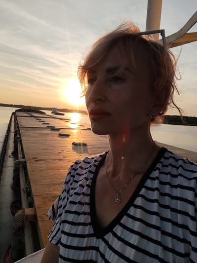 A medium shot captures a woman with short, reddish-blonde hair and a striped V-neck shirt standing on the deck of a barge at sunset. The woman is looking to the left, her face partially obscured by the warm glow of the setting sun. Her lips are painted red, and she wears a heart-shaped earring and a silver necklace with a crown pendant. Behind her, the wide, flat deck of the barge stretches towards the horizon, dotted with dark, rectangular hatch covers. A calm river reflects the golden hues of the sky, and a small vessel is visible in the distance. The scene takes place on the Volga River near the village of Shestovo, Russia.