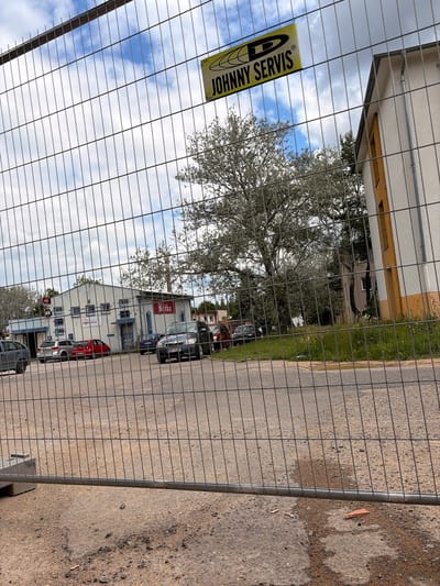 The image shows a construction site with a temporary wire mesh fence in the foreground, obscuring the view of a building and cars parked in front of it. A yellow sign with black lettering that reads "JOHNNY SERVIS" is attached to the fence. Behind the fence, a light-colored building with blue trim and windows is visible. There are several cars parked in front of the building, including a dark SUV, a red car, and a silver car. The building appears to be a commercial establishment, with signage that includes the word "Běčko" in red lettering. A large tree with sparse foliage stands to the left of the building. The sky is cloudy with patches of blue. The scene appears to be a street or parking lot in an urban or suburban area. The location is confirmed to be Příbram, Czechia, based on the context provided.