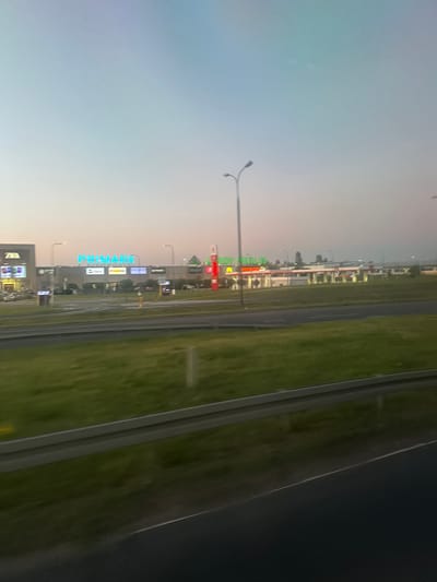 The image depicts a view of a shopping center and a gas station on the outskirts of Lublin, Poland, captured during dusk. The primary structure visible is a large retail complex with prominent signage for "Primark" illuminated in bright blue neon. To the left of Primark, another building with "ZARA" signage is partially visible. Several other smaller signs for various stores, such as "TARA" and "SEPHORA," are also discernible. In the mid-ground, a gas station with red and white pumps and canopy is illuminated. Streetlights are present throughout the scene, both within the shopping center complex and along the adjacent road. The foreground is dominated by a grassy median strip with a guardrail, separating the viewer's perspective from the road and the commercial area. The sky transitions from a pale blue at the top to a warmer, hazy orange near the horizon, indicating either sunset or sunrise. The image is slightly blurred, suggesting it was taken from a moving vehicle.