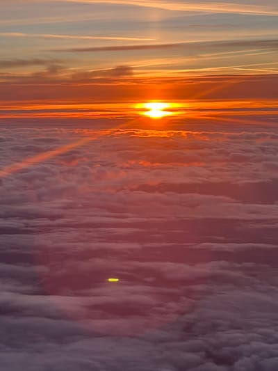 The image depicts a vibrant sunset viewed from above a dense cloud layer. The sun's intense orange light pierces through the clouds, creating a warm glow. The thick, undulating cloudscape stretches across the majority of the frame, casting deep purple and gray shadows. The photograph appears to be taken from an elevated position, potentially an airplane. There is no evidence to suggest that this image was taken in Steenbergen, Netherlands.