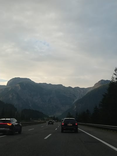 The image depicts a view from a car on a highway in Flachauwinkl, Austria, with a mountain range in the background. The sky is overcast with clouds. Several cars are visible on the highway, including a black SUV in the foreground on the left with its taillights illuminated, and another black SUV further ahead on the right. Trees and mountains form the backdrop, with the mountains appearing dark and rugged, partially obscured by clouds. The highway has multiple lanes with white markings. The scene appears to be during the daytime, possibly late afternoon or early evening, given the lighting conditions.