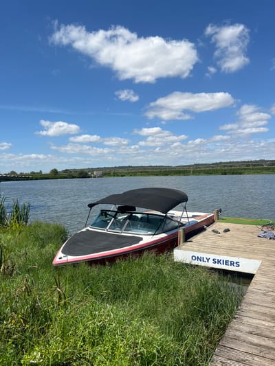 The image displays a red and white pleasure boat, equipped with a black bimini top and a black bow cover, docked at a wooden pier. The boat's bow points towards the overgrown grassy bank on the left, where tall green grass and reeds are abundant. A white sign with blue text reading "ONLY SKIERS" is affixed to the side of the pier facing the water. On the pier's wooden planks, a pair of black flip-flops and a folded grey textile item are visible. The boat is moored in a calm body of water with slight ripples, reflecting the blue sky and scattered white cumulus clouds above. In the mid-ground across the water, green vegetation lines the distant bank, where a large, curved irrigation system and a small, light-colored building are discernible. A bridge structure spans the water in the far left background. The scene is located in Coruche, Portugal, under bright daylight conditions. No individuals are present.