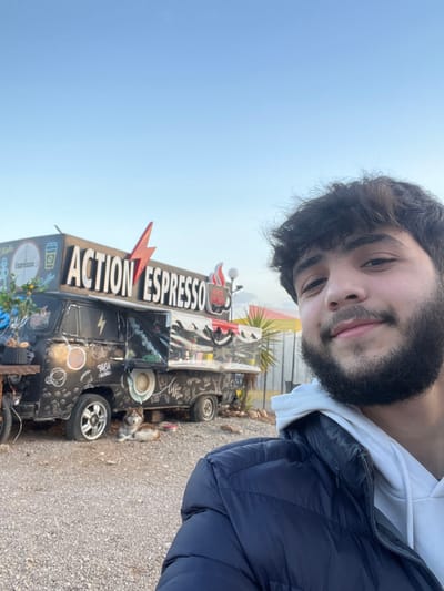 A young adult male, with dark, medium-length hair and a full dark beard, is depicted in a self-portrait. He is wearing a white hoodie beneath a dark blue puffer jacket and exhibits a slight smile. The background reveals a customized black food truck, presumably located in Qudsaiya, Syria, serving as a mobile coffee stand.

The food truck, likely a converted van, features a prominent sign on its roof that reads "ACTION ESPRESSO" in white capital letters, accented by a red lightning bolt graphic between the words. The van's exterior is extensively decorated with white line art depicting coffee cups, coffee beans, and phrases such as "Fresh Beans" and "Coffee." An open serving hatch is visible on the vehicle's right side (viewer's left). A string of decorative lights is attached to the side of the truck. To the left of the van, a small plant with visible orange fruits, possibly citrus, is situated in a dark container.

On the ground in front of the truck's left front wheel (viewer's right), a light-colored dog, resembling a Husky or Malamute with brown and white fur, lies on its stomach. The ground surface consists of light-colored gravel or small pebbles. The sky above is clear and light blue. In the distant background, behind the food truck, a tall, palm-like plant and a segment of a white corrugated metal fence or structure are discernible.