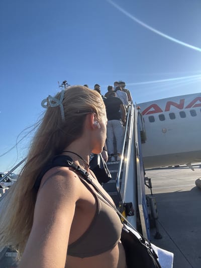A low-angle, full shot captures people boarding an airplane via a mobile staircase under a clear blue sky. In the foreground, a young woman with long blonde hair, secured with a light blue hair clip, faces away from the camera, looking towards the airplane. She is wearing a dark gray halter-top bra and a black backpack is slung over her shoulder. Her right ear is adorned with a white wireless earbud.
The staircase, constructed from metal with yellow and black striped safety markings, leads up to the airplane's entrance. Several individuals, dressed casually in shirts and pants, are ascending the stairs in a single file line, their backs to the camera.
The airplane, a commercial passenger jet, is predominantly white with a red logo spelling "ANA" visible on its fuselage. The aircraft is parked on an asphalt tarmac. In the background, a sliver of another vehicle, possibly a service truck, is visible near the left edge of the frame. The bright sunlight casts long shadows across the tarmac, indicating it is likely late afternoon or early morning. The location is the city of Pendik, Turkey.