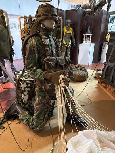 The image depicts an indoor military museum exhibit located in Praia do Ribatejo, Portugal. In the foreground, a mannequin is kneeling on a light-tan floor mat, dressed in a green and brown camouflage military uniform, helmet with a camouflage cover, olive-drab gloves, and black combat boots. It wears a full military harness with a large backpack and a chest-mounted pouch. Its gloved hands are holding multiple white parachute suspension lines that extend to a section of white parachute canopy fabric laid out on the floor, simulating the act of packing a parachute. To the mannequin's left, a camouflaged box or crate with attached cords rests on the floor.
To the left of the central display, a portion of an olive-drab parachute container with "EFA 500" text is visible. In the mid-ground, another standing mannequin wears an olive-drab flight suit with a prominent yellow harness, helmet, and goggles. Further to the right and elevated, a third mannequin, dressed in camouflage, is suspended horizontally, depicting a parachutist in flight or descent. Between the standing and suspended mannequins, a white display pedestal holds a miniature gray architectural structure, possibly a castle, atop a wooden base, alongside a small red object. The museum environment features light-colored walls, dark brown partition walls, framed pictures on the background walls, and various packed parachute bags on the floor. The flooring transitions from a tiled section in the far left background to a consistent light-tan mat or carpet in the main display area.