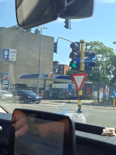 The image shows a view from inside a vehicle, looking out towards a Q8 gas station and traffic lights in Boom, Belgium. A blue Renault car is stopped at a red light, with a silver car to its left. The traffic light shows a green signal, indicating that the light has just changed. The gas station has multiple pumps and a "shop & go" convenience store. Trees and buildings are visible in the background. The scene is captured on a sunny day.