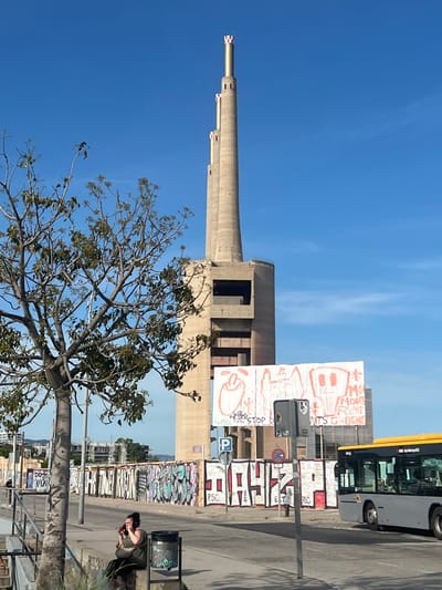 In Sant Adrià de Besòs, Spain, a tall, Brutalist concrete tower with three smokestacks, each topped with red and white checkerboard patterns, dominates the skyline under a clear blue sky. In the foreground, a young woman with dark hair tied up sits on a concrete ledge, holding a phone to her ear and appears to be talking on it. She is wearing a dark green sleeveless top and a dark crossbody bag. Beside her is a black trash receptacle. A bare-branched tree with a few green leaves is positioned to the left of the frame. A large white billboard with a red, crudely drawn, abstract mural is affixed to the base of the tower, with graffiti covering a low wall in front of it. A yellow-striped city bus is visible driving on the right side of the street. The street is lined with buildings and more graffiti.