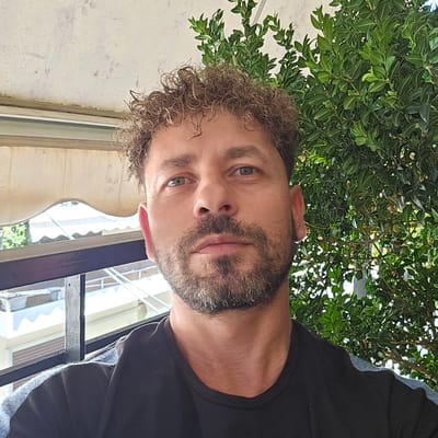 The image is a close-up selfie of a man with curly brown hair and a beard, taken outdoors. He is wearing a black t-shirt. Behind him, there is a dense green hedge, a railing, and a blurred background that appears to be an urban environment with buildings and awnings. The lighting suggests it is daytime. The man's facial expression is neutral. The image was captured in Ilion, Greece.