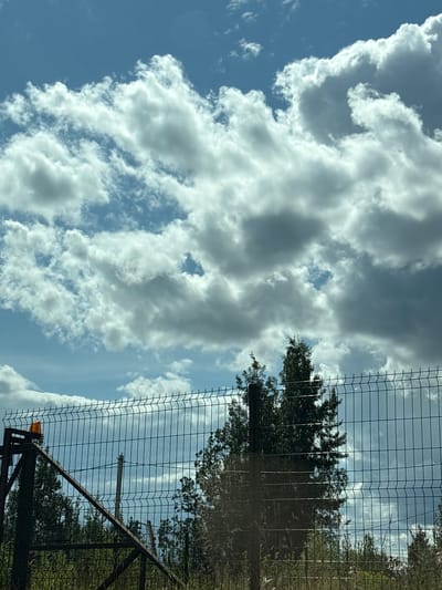 The image depicts a clear sky with scattered clouds. A metal fence with an orange warning light on a post runs across the lower half of the frame, partially obscuring trees and vegetation behind it. The scene appears to be outdoors, possibly near a construction site or a fenced-off area. There are no people or discernible actions taking place. The location is stated to be in the city of Imeni Sverdlova, Russia.