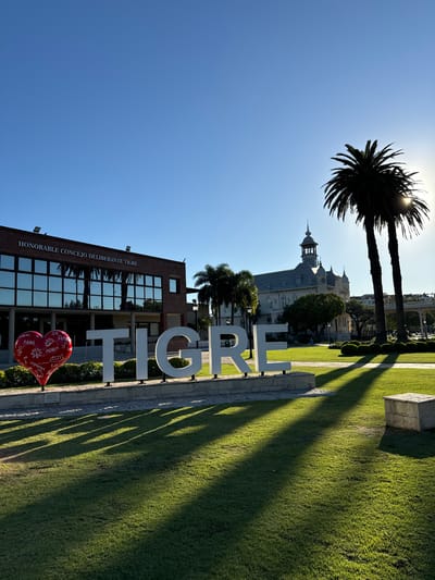 The image depicts a sunny day in Tigre, Argentina, with the white "TIGRE" sign prominently displayed in front of the Honorable Deliberative Council of Tigre building. A red heart-shaped object stands near the base of the sign. In the background, the Museo de Arte Tigre building is visible along with two palm trees that reach into the sky. The photo is taken from a low angle. The sun is positioned behind the palm trees, creating a bright glare and casting long shadows across the green grass. The image is rotated 90 degrees counter-clockwise.