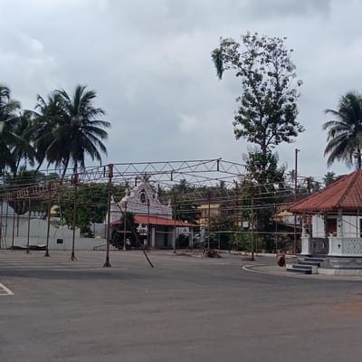 The image depicts an outdoor scene in Marna, India. A large, unfinished metal framework dominates the foreground, suggesting construction or preparation for an event. Palm trees are visible in the background, along with a building with a white facade and a red-tiled roof. To the right is a small, white gazebo with steps. The ground is a paved area, possibly a parking lot or public square, marked with white lines. The sky is overcast, suggesting a cloudy day. A single individual is visible near the gazebo.