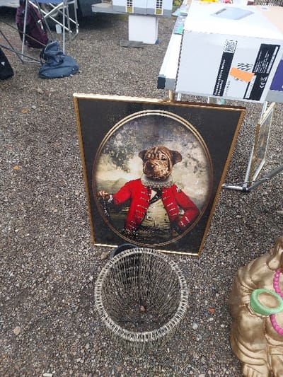 The scene, located in Eckbolsheim, France, depicts an assortment of objects arranged on a gravel surface. A prominent rectangular picture frame, gold-toned with a dark inner border, stands upright, displaying an oval portrait. The portrait depicts the head of a dog, possibly a Shar-Pei breed, atop a human body dressed in a red, collared jacket with white ruffles at the neck and cuffs, a white shirt, and a dark, possibly tricorn, hat. The background of the portrait features a muted landscape. Adjacent to the framed artwork, on the gravel, is a circular, open-top basket constructed from woven wire or thin metal strips. To the right of the basket, partially visible, is a golden statue, consistent with a Buddha figure, adorned with a pink beaded necklace and holding a green, cup-like object. In the upper left background, a dark blue duffel bag or backpack rests on the gravel near the exposed metallic frame of a folding chair. In the upper right background, a white cardboard box with visible black and orange labels sits on a metallic display stand, positioned next to a miniature architectural model of a house featuring white walls and yellow window frames. The entire ground plane is composed of small, irregular gray and brown gravel stones.