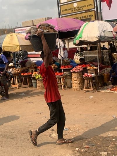 This image depicts an active outdoor market scene in Otuocha, Anambra State, Nigeria, under daylight conditions with an overcast sky allowing for direct sun patches. The ground is unpaved dirt with scattered small debris.
In the mid-ground, a male individual walks from right to left, facing away from the viewer's immediate right. He carries a large, dark-colored plastic basin filled with organic material, including what appears to be soil or refuse mixed with plastic bags, balanced on his head. His arms are raised, hands holding the sides of the basin for stability. He is wearing a red short-sleeved t-shirt, dark-colored long pants, and dark-colored flip-flops. His shadow extends diagonally behind him on the dusty ground.
The background is filled with various market stalls. Produce is displayed on numerous wooden stools and woven baskets, including piles of red tomatoes in metal trays, green fruits in a smaller tray, brown root vegetables, and light-colored cuts of meat or fish on flat trays. To the far right, small red-skinned vegetables are visible on a ground mat. Market umbrellas, faded in color (yellow, pink, and green/white), provide overhead shade. Parts of other individuals are visible among the stalls, including one wearing a blue shirt near a motorcycle on the far left, and another person under a pink umbrella.
Key identifying signs are visible in the background: a yellow sign reads "GOVERNMENT OF ANAMBRA STATE OF NIGERIA", and partially below it, "PRO... OF SHELL ROAD," and "LOCAL GOVERNMENT ARE". Another sign on a building under construction reads "PROJECT DESCRIBING BUILDING OF OTU...". A red vehicle is partially visible behind the stalls on the right.