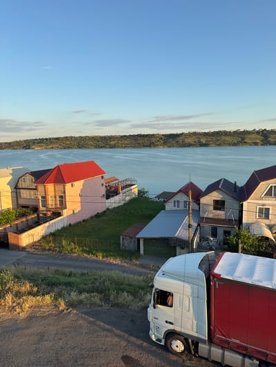 The image shows a waterfront community in Yelyzavetivka, Ukraine, bathed in the warm light of late afternoon or early morning. In the foreground, a white semi-truck with a red trailer is parked on a gravel road, casting a long shadow. The truck has a DAF logo visible. Behind the truck, a collection of houses with red and gray roofs lines the shore of a calm body of water, likely the Dnieper River. The houses vary in architectural style, with some featuring balconies and air conditioning units. The landscape is gently sloped, covered in green grass and scattered trees. In the background, the opposite bank of the river is visible, characterized by a tree-covered hillside dotted with more houses. The sky is clear blue with a few wispy clouds.