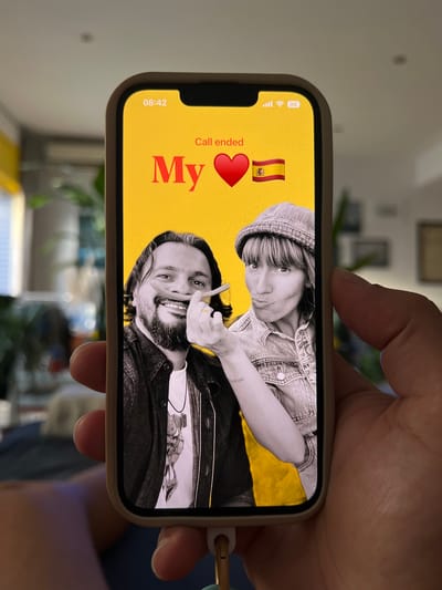 The image shows a hand holding a smartphone displaying a video call screen. The screen is yellow with red text that reads "Call ended My ❤️🇪🇸". On the screen, two individuals are visible. On the left, a man with dark hair and a beard is smiling and holding what appears to be a cigarette or thin stick to his mouth. He is wearing a dark jacket over a white t-shirt with a necklace. On the right, a woman with short, layered hair and a bucket hat is making a kissing face. She is wearing a denim jacket. The background of the video call screen is a solid yellow. The background outside the phone is blurred but appears to be an indoor setting with hints of furniture and plants. The time displayed on the phone is 08:42. There is no discernible information to suggest the location is in Mollet Del Vallès, Spain.