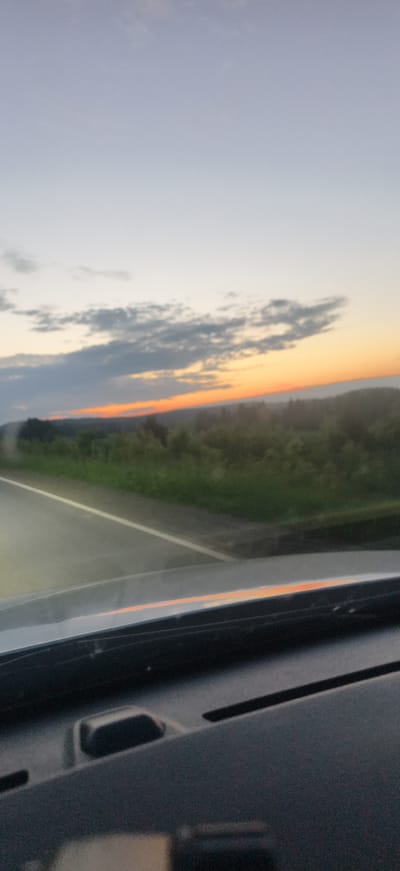 The image depicts a view from inside a vehicle, looking out at a rural landscape at dusk. A road with a white line is visible in the foreground, leading into a field of green vegetation and trees. The sky is a gradient of pale blue to orange, with scattered clouds. The dashboard of the car is visible in the lower portion of the image. The scene is located in the village of Забегалово, Russia.