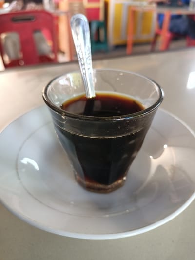 A close-up, eye-level shot captures a clear glass filled with dark coffee, resting on a white saucer. A silver spoon is partially submerged in the coffee. The background is softly blurred, showing glimpses of red and yellow chairs, and a colorful table, suggesting an indoor seating area of a cafe or eatery, possibly in Pulokiton, Indonesia, given the context. The lighting appears natural, with light reflecting off the glass, saucer, and spoon. The overall impression is that of a simple moment at a cafe.