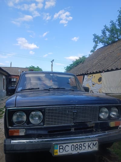 The image shows the front of a dark blue car, likely in a rural or suburban setting in Pishchanka, Ukraine. The car is angled towards the viewer, with its grille and headlights prominently displayed. A license plate with the characters "BC 0885 KM" is visible on the front of the car, along with a Ukrainian flag emblem.

The background features a rustic roof, possibly part of a house or shed, with some greenery and trees to the left. The sky is a clear blue with scattered white clouds, suggesting it is daytime with fair weather. The overall scene appears to be a still, perhaps candid, shot of the car. There are no people visible in the image. The car seems to be parked. The image has a somewhat aged or low-resolution quality, and is oriented vertically.