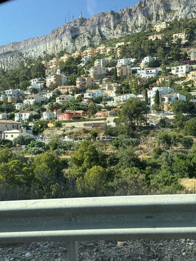 This media file depicts a steep, rocky cliff face in Calp, Spain, dotted with a dense cluster of white and beige houses and lush green trees. The houses are built into the hillside, with many featuring terraces and balconies, suggesting a Mediterranean architectural style. The scene is bathed in bright sunlight, indicating it is daytime, likely mid-morning or early afternoon, with clear weather. The overall impression is one of a vibrant, sun-drenched coastal town clinging to a dramatic natural landscape. A gray metal guardrail runs vertically through the right side of the image, with visible gravel and rough terrain to its right. The orientation of the image is rotated 90 degrees counter-clockwise, making the cliff face appear to be on the left and the houses cascading down the right.