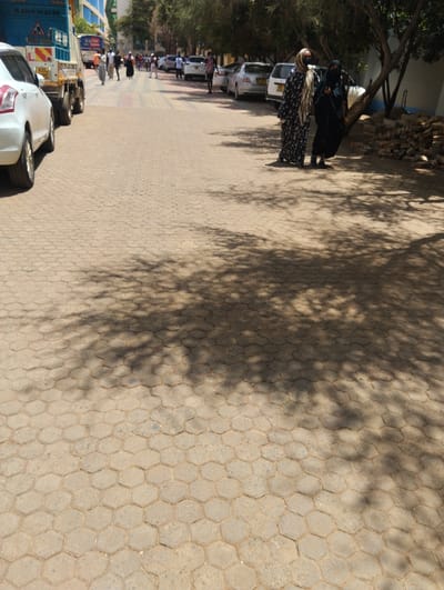 The image depicts an outdoor street scene in Hospital ward, Kenya, under bright daylight. The ground is paved with hexagonal interlocking concrete blocks, exhibiting wear. Strong, dappled shadows from unseen trees cover the right side of the paved surface.
In the foreground on the right, two women are standing on the paved surface near a tree. The woman on the left wears a long, black and white patterned garment and a light-colored head covering that partially obscures her face. The woman on the right wears a solid black long garment and a black niqab, revealing only her eyes. To their immediate right, under the tree, is a small pile of reddish-brown bricks or construction debris.
In the midground, further down the paved street, a group of approximately eight to ten individuals are walking away from the camera. Vehicles are parked or stationary along both sides of the street. On the far left, a white hatchback car, partially visible, has a license plate with "KBC". Behind it, a light blue flatbed truck with low side rails displays the license plate "KBC 117E" and a blue sign with "070X 15 32 XX" and a triangular hazard symbol. Multiple other light-colored sedan cars are visible parked along the right side of the street in the distance. Tall buildings are present in the far background on the left. No direct interactions are evident between the two foreground women and the individuals in the midground.