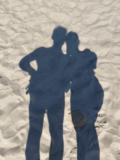 Two shadows of people are cast on the sandy ground. One shadow, on the left, appears to be that of a male figure with his left hand on his hip. The shadow on the right, possibly of a female figure, is positioned slightly behind and to the right of the first. The figures' heads are close together, suggesting they are interacting. A small, light-colored rock is visible on the sand between the shadows of their legs. The environment is a beach with textured sand, indicating natural light and possibly a sunny day. The specific location within the city of Yasnyska, Ukraine, is not discernible from the image itself.