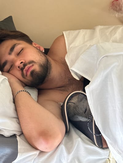 In Bliny, Russia, a man is sleeping on a bed. He has dark hair and a beard and is wearing a silver hoop earring. He is lying on his side with his left arm under his head, which is resting on a white pillow. A white sheet is pulled up to his chest. A black and brown zippered bag with a mesh top is next to his right arm. The background is a plain, light-colored wall.