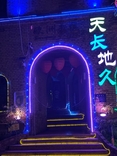 The image depicts a building facade in Baqiao, China, adorned with colorful neon signs. The signs display various messages, including "Tianchangdijiu" (天长地久), which translates to "eternity," "Guangpandong" (光盘行动), which translates to "Clean Plate Campaign," and "Maidong" (脉动), a Chinese brand name for a sports drink, and signs with safety messages. The building is constructed of brick and features an arched entryway highlighted by a purple and blue neon outline. A line of yellow lights vertically extends from the structure to the right of the arched entrance. Surveillance cameras and electrical wiring are visible, adding to the urban aesthetic of the scene. A couple of security and caution signs are visible at the lower right part of the image.