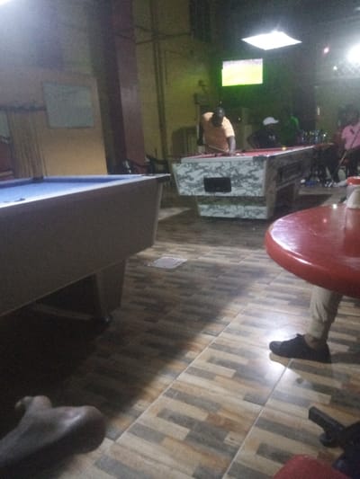 The image depicts an indoor recreational establishment located in Majengo ward, Kenya. The scene is dimly lit, featuring at least two pool tables and several individuals.

In the midground, an adult male, wearing an orange short-sleeved shirt, is positioned over a pool table with a red felt surface and a white and green camouflage-patterned base, appearing to execute a shot. Behind him and slightly to the right, a second male, clad in a dark top and a white cap, is visible near the same pool table. Further to the right and in the background, a third individual, wearing a pink shirt, is partially visible, seated or standing near a table with multiple bottles. A flat-screen television is mounted on the far wall, displaying a brightly illuminated image. A square ceiling light fixture provides overhead illumination.

In the foreground on the left, a portion of a pool table with a blue felt surface is visible. On the extreme lower left, a bare foot is positioned on the tiled floor. On the right side of the foreground, a red circular table is partially visible, with a white cylindrical container on its surface. The lower legs and feet of an individual, wearing light-colored trousers and black shoes, are visible beside this red table. The floor throughout the establishment is covered with rectangular ceramic tiles in varying shades of brown, beige, and gray, arranged in a staggered pattern. The walls appear to be a dull yellow or beige, with a dark vertical column visible towards the center-left. A plain white board is mounted on the left wall.