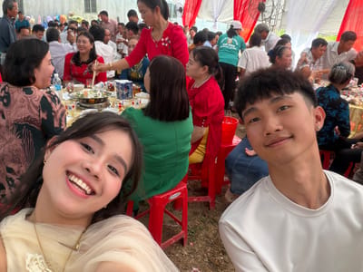 This image depicts a vibrant outdoor gathering, likely a communal meal or celebration, in Tay Son Commune, Vietnam. In the foreground, a man and a woman are taking a selfie. The woman on the right is smiling broadly with an open-mouthed expression, revealing her teeth, while the man on the left has a more subtle smile. Both appear to be in good spirits.
Behind the two main subjects, numerous people are seated at multiple tables arranged under temporary white and red fabric canopies. The tables are laden with food, including what appears to be a hot pot in the center of one table, surrounded by various dishes, cups, and bottles of water. Pepsi cans are also visible on the tables. Attendees are engaged in eating, conversing, and socializing. The chairs are predominantly red plastic, with at least one bearing the visible brand name "DUY TAN".
The event is held outdoors on a patch of ground that looks like dirt or sparse grass. A large fan is visible towards the bottom of the image, suggesting a warm climate. The natural lighting indicates that the event is taking place during the daytime. Visible text on a green t-shirt in the background includes "CỔ HOÁ", "CO HOA", "CHUYÊN VIỆT", and a partial phone number. The overall scene conveys a lively and communal atmosphere.