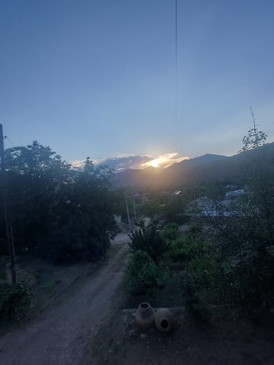 The image depicts a rural landscape at sunset in the vicinity of Areni, Armenia. The sky is a gradient of blue, transitioning to lighter hues near the horizon where the sun is setting behind distant mountains. The sun's rays break through clouds, casting a warm glow on the scene. In the foreground, dark green trees dominate the left side of the frame, obscuring a dirt road that winds through the landscape. Several houses with visible roofs are scattered throughout the middle ground, nestled among trees and vegetation. Two clay pots are placed on the ground in the lower right corner of the image. A thin, dark line, likely a cable or wire, extends vertically from the top of the frame towards the horizon. The overall atmosphere is serene and quiet, characteristic of a rural evening.
