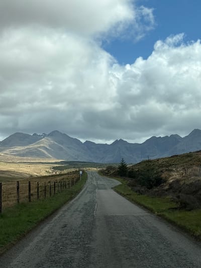 A road in the Isle of Skye, United Kingdom, is depicted under a cloudy sky. The road runs vertically along the right side of the image. To the left of the road, there's a grassy area, then a field, and beyond that, a mountain range. The sky is predominantly filled with grey clouds, with a small patch of blue sky visible in the upper left corner.