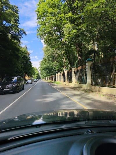 The media file shows a view from inside a car driving down a tree-lined road. The scene is set in Arkhangel'skoe, Russia, on a bright, sunny day with a few clouds in the blue sky. Tall trees with lush green foliage flank both sides of the road, casting shadows on the asphalt. On the right side of the road, there is an ornate black metal fence with decorative stone pillars. Several cars are visible on the road ahead, traveling in the same direction. The car in the foreground on the left is a black Mercedes-Benz Vito with the license plate "K230OO 774". The dashboard of the car from which the photo is taken is visible in the lower part of the frame, with reflections of the trees and sky on the windshield. The overall atmosphere is peaceful and serene, suggesting a leisurely drive through a scenic area. The time of day appears to be mid-morning or early afternoon, given the bright sunlight and the quality of the shadows.
