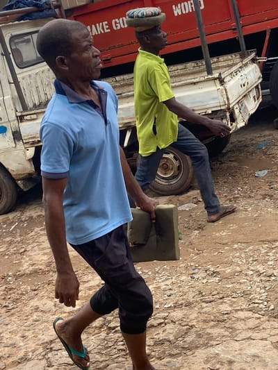 In Ogbunike, Nigeria, two men are walking in a dirt-covered area next to a truck. The man in the foreground, wearing a light blue polo shirt and dark pants, is carrying a green cushion. He is walking towards the left of the frame, with his head turned to the right. The second man, in a neon yellow shirt and dark pants, is walking behind him, carrying a cushion on his head. The truck is white with red accents and appears to be old and worn. The ground is uneven and rocky, with some loose gravel. The overall scene suggests a busy, working environment.