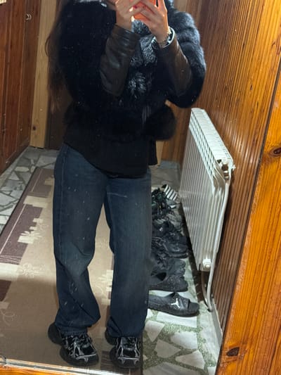 A person wearing a black fur-trimmed jacket, dark jeans, and black boots stands inside a room with wooden walls and ceiling. The person is holding a red object, possibly a phone. Above the person, there is a white radiator hanging from the ceiling. Below the radiator, there are several pairs of black shoes or sandals. The floor is covered with a patterned rug. The location is likely inside a house, but the city of Rudartsi, Bulgaria, cannot be confirmed from the image alone.