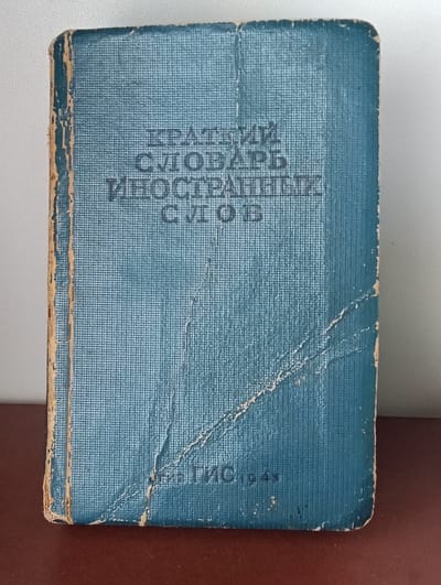 A closed, antique, blue hardcover book with worn edges and a textured cover is displayed. The book is positioned vertically on a dark brown surface. The title, written in dark, capital letters, is visible on the cover. It reads "КРАТКИЙ СЛОВАРЬ ИНОСТРАННЫХ СЛОВ", which translates to "A Brief Dictionary of Foreign Words". In the bottom right corner of the book cover, the inscription "ТИС. 1943" is also visible. The background is a plain, light-colored wall. The book is the sole subject of the image.