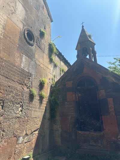The image shows a section of the Bjni Fortress in Bjni, Armenia, featuring ancient stone structures bathed in sunlight under a clear blue sky. On the left, a large, weathered stone wall is covered in carved inscriptions. A circular window with decorative stonework is visible on this wall. To the right, a smaller stone building with a pointed bell tower stands, topped with a cross. The bell tower has an arched entrance and a dark, possibly wooden or metal door. Green vegetation grows in patches on the stone walls, suggesting age and weathering. The overall scene depicts historical architecture and an outdoor environment with strong natural lighting.